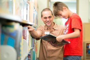 Male librarian with goatee squatting next to a male child in the library looking at the book Hip Hop Dog.